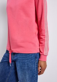 Felpa rosa con orlo a coulisse, caratterizzata da strisce bianche lungo le maniche. Il materiale sembra morbido, abbinato a jeans in denim blu.