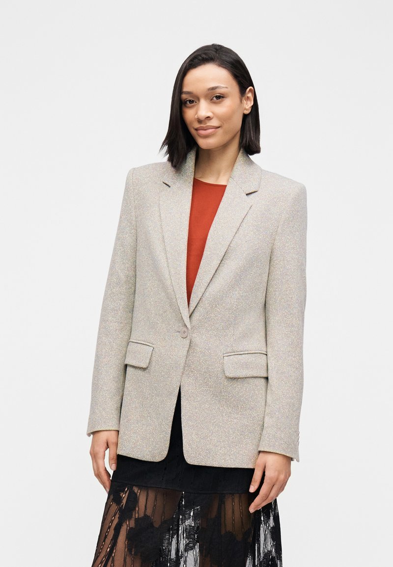 Lichtgrijze getextureerde blazer met ingesneden revers, knoopsluiting aan de voorkant en twee zijzakken, gedragen over een rood topje en een zwarte kanten rok.