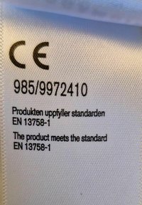 Weißes Stoffetikett mit schwarzer Schrift, das das CE-Zeichen, Produktnummern und Compliance-Informationen auf Englisch und Schwedisch anzeigt.