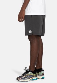 Gray shorts with a fabric tag featuring blue and white logo. Worn with multicolored athletic shoes and a white T-shirt.