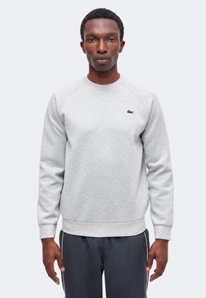 Homme portant un sweat-shirt gris clair à manches longues avec un petit logo vert sur la poitrine et un pantalon foncé avec des bandes blanches sur les côtés.