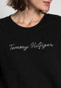 Czarna bluza z okrągłym dekoltem, zdobiona srebrnym haftem z napisem "Tommy Hilfiger" na przodzie. Gładka tekstura materiału.