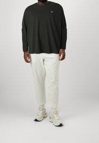 Černé tričko s dlouhým rukávem a kulatým výstřihem, bílé kalhoty a béžové tenisky. Výrazné logo na hrudi a světle šedé pozadí.