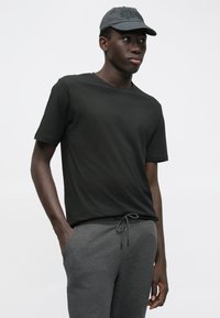 Jonge man in een zwart t-shirt, grijze sweatpants met trekkoord en een donkergrijze pet, die met één hand in zijn zak staat tegen een witte achtergrond.