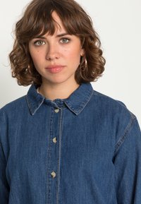 Camisa de ganga com gola, de cor azul claro, apresentando fecho com botões na frente e detalhe de costura visível nos ombros.