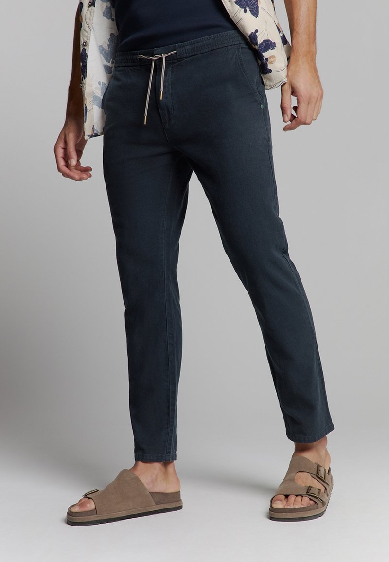 Heren donkerblauwe katoenen broeken met een trekkoord in de taille, tapered pijpen en subtiele textuur; gecombineerd met bruine open teensandalen.
