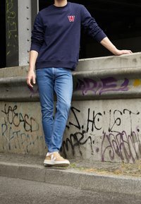 Marinblå sweatshirt med röd logotyp, blå slim-fit jeans och beige sneakers. Bakgrunden har graffiti på en betongstruktur.