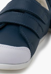 Chaussure bleu marine avec un devant texturé, un bout en caoutchouc blanc et deux bandes Velcro sur le dessus. Matériau en cuir lisse.