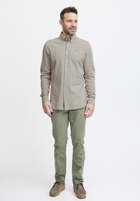 Chemise beige à manches longues avec des boutons et un petit logo, associée à un pantalon slim-fit vert olive et des chaussures en daim marron.