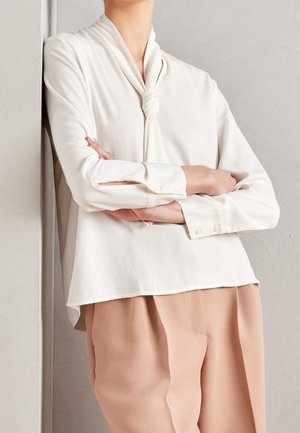 Personne les bras croisés portant un chemisier crème avec un col noué et un pantalon beige à jambes larges sur un fond uni.