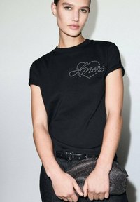 Crna majica s kratkim rukavima, ukrašena dizajnom srca od rhinestona na prsima. Model drži teksturiranu, svjetlucavu torbicu.