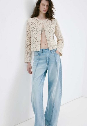 Mujer joven con cabello rizado que lleva un cárdigan de crochet crema sobre el torso desnudo y vaqueros azul claro de talle alto y pierna ancha.