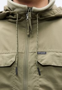Φούτερ μπουφάν σε ελαιώδες πράσινο με κουκούλα και φερμουάρ, δύο μπροστινές τσέπες, απαλή φλις επένδυση, ρυθμιζόμενα κορδόνια και λογότυπο Columbia στο στήθος.