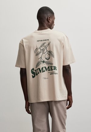 Homme de dos portant un t-shirt beige avec un imprimé botanique noir et le texte « Never Ending Summer Filled with Adventure » dans le dos.