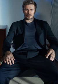 Fekete zakó sötétkék ing felett, ülő férfi látható tetoválásokkal, rövid hajjal és laza testtartással egy világos háttér előtt.