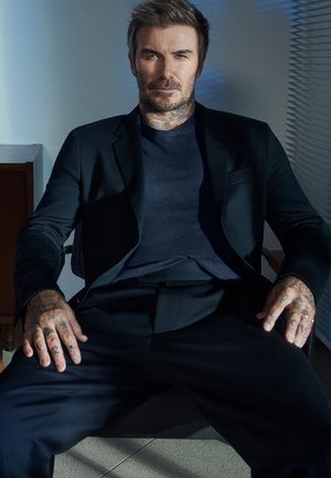 Zwarte blazer over een marineblauw shirt, zittende man met zichtbare tatoeages, kort haar en een ontspannen houding tegen een lichte achtergrond.