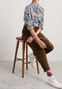 Camisa con estampado floral en blanco, azul y rojo; pantalones de pana marrones; zapatillas blancas; sentado en un taburete de madera contra un fondo neutro.