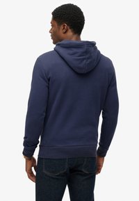 Marineblauwe hoodie met een zachte textuur, met een grote capuchon, lange mouwen en geribde manchetten. Draagt hij met donkere spijkerbroeken.
