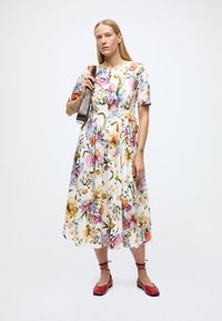 Bloemen midi-jurk met korte mouwen, witte achtergrond, kleurrijke bloemenprint. Model houdt een bruine tas vast, draagt rode ballerina's met enkelstrikjes.