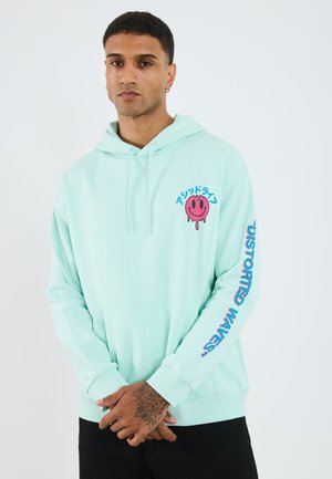 Myntegrønn hettegenser med et frontlogo som viser et rosa smilende ansikt. Blå tekst nedover ermet sier "DISTORTED WAVES." Mykt stoff og snor.