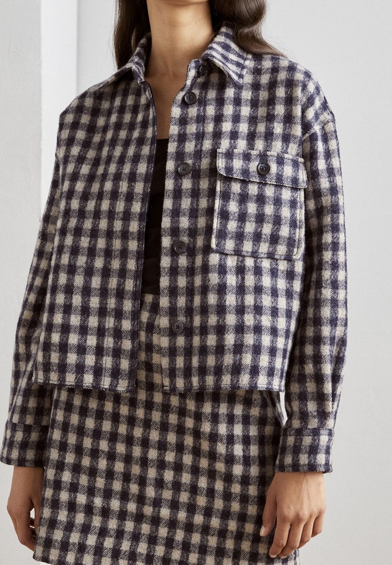 Femme portant une veste courte à carreaux violets et blancs avec des boutons, assortie à une jupe, debout devant un fond clair uni.