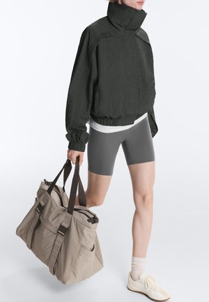 Person trägt eine dunkle, übergroße Jacke, graue Radhosen, weiße Socken und Turnschuhe, und trägt eine große beige Sporttasche mit dunklen Trägern.