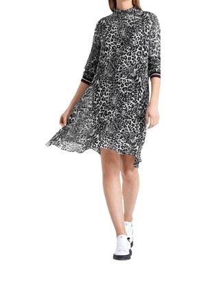 Woman wearing a black and white leopard print knee-length dress with three-quarter sleeves and white sneakers, standing against a white background.