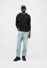 Black knitted hoodie with a relaxed fit, light blue straight-leg jeans, and black sneakers, complemented by a gray cap. Minimalist design.
