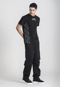 Mustat t-paidat, joissa on logo rinnassa ja kuvioidut sivut; yhdistetty mustiin cargo-housuihin, joissa on useita taskuja ja mustiin lenkkareihin.