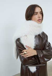 Donna che indossa un cappotto di pelle marrone scuro, avvolta in una sciarpa bianca e morbida, e con un grande anello d'oro sulla mano sinistra, che guarda di lato.