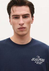 Navy blue crew neck t-shirt made of cotton, featuring white embroidered text on the chest, against a plain background.