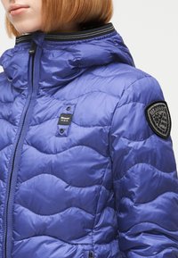 Gros plan d'une personne portant une doudoune matelassée bleue avec une capuche, un col bordé noir et blanc, et des écussons de marque sur la poitrine et la manche.