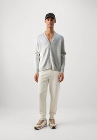 Graue Strickjacke mit V-Ausschnitt und Knopfverschluss, kombiniert mit cremefarbenen Jogginghosen und beigen Turnschuhen. Das Modell trägt eine schwarze Mütze.