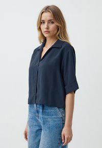 Blusa corta a maniche corte di un blu navy scuro con una vestibilità ampia, colletto a bavero e chiusura frontale con bottoni. Tessuto morbido e liscio con una leggera lucentezza.