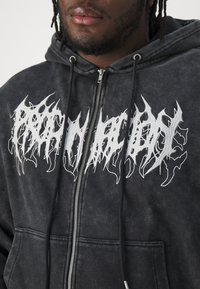 Juodas zip-up bliuzonas su nusidėvėjusiu tekstūru, turintis išraiškingą baltą grafiką logotipą krūtinėje ir raištelius ant gobtuvo.