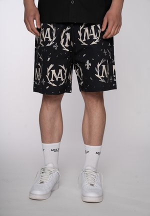Man draagt zwarte korte broek met patroon en witte logo's, witte sportsokken met tekst en witte sneakers, staand tegen een effen achtergrond.