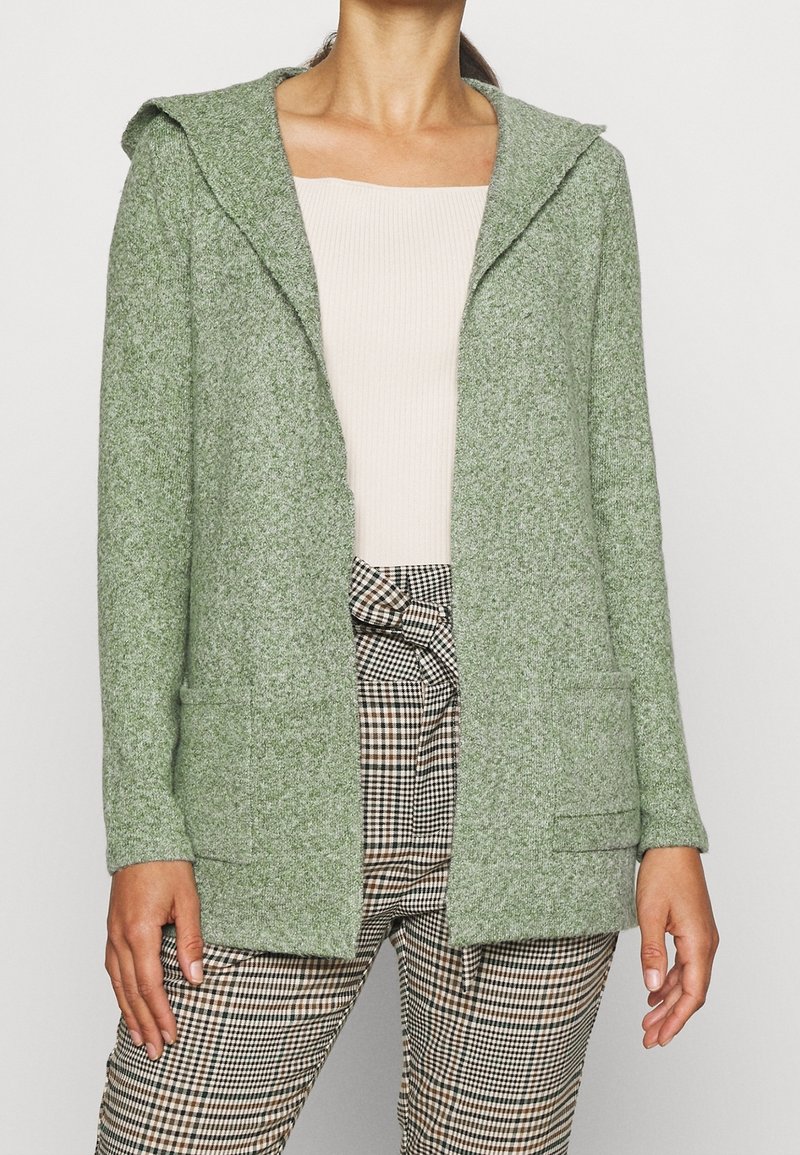 Femme portant un cardigan à capuche vert clair, un haut côtelé crème et un pantalon à carreaux marron et noir taille haute avec une ceinture nouée.