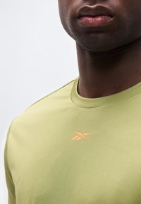 Κοντινό πλάνο ενός ατόμου που φοράει ανοιχτό πράσινο πουκάμισο με πορτοκαλί λογότυπο Reebok στο κέντρο του στήθους, με ορατά τα χείλη και το κάτω μέρος του προσώπου.