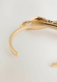 Goldfarbener Armreif mit einer glatten, polierten Oberfläche und einem abstrakten Design, das subtile Verzierungen und eine fließende Form aufweist.