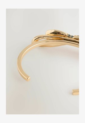 Goldfarbener Armreif mit einer glatten, polierten Oberfläche und einem abstrakten Design, das subtile Verzierungen und eine fließende Form aufweist.