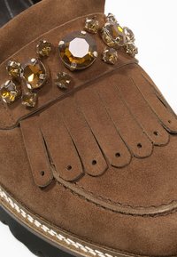 Mocassin en daim marron avec détail à franges et ornements de pierres précieuses facettées de couleur ambre sur la partie supérieure.