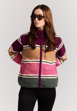 Vrouw met een zonnebril en een meerkleurige fleecejas met horizontale strepen in paars, beige, roze en groen, staand met handen achter de rug.