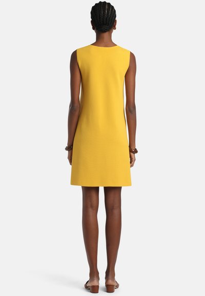 Femeie cu părul împletit, în picioare, purtând o rochie galbenă fără mâneci și sandale maro cu toc, cu fața către spate, pe un fundal alb.