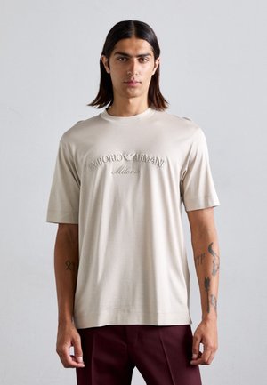 Beige cotton t-shirt with short sleeves, featuring embossed "Emporio Armani Milano" logo. Smooth texture with a relaxed fit.