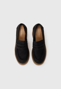 Mocassins en daim noir avec un bout arrondi, dotés d'une sangle décorative et d'une semelle en caoutchouc brun clair. Texture lisse avec des accents réduits.