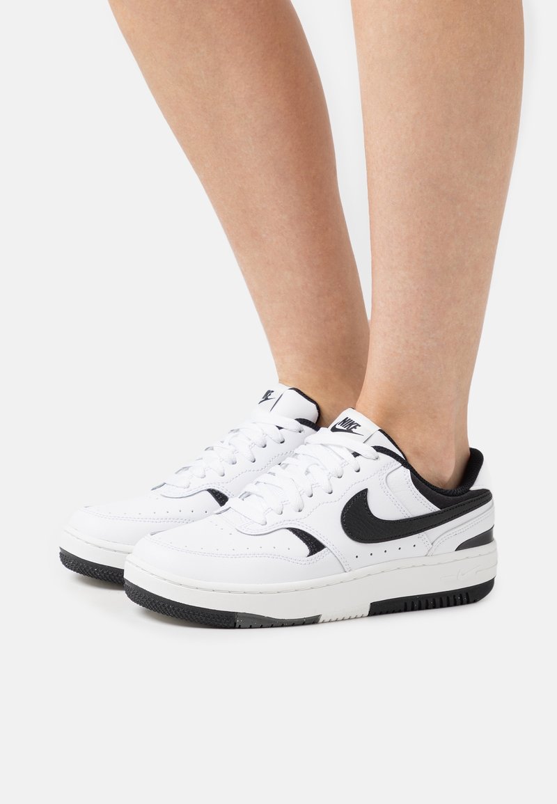 Baskets en cuir blanc avec des accents noirs, featuring un design basse, bout rond et semelle en caoutchouc texturé. Lacets et détails perforés visibles.