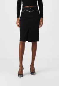 Mujer con una falda negra a la altura de la rodilla con abertura frontal, cinturón de cadena, camiseta negra de mangas largas y zapatos de tacón alto negros con puntas metálicas.