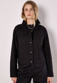 Czarna kurtka jeansowa z zapięciem na guziki, dwoma kieszeniami na piersi i usztywnionym kołnierzem. Posiada pionowe przeszycia i krótki krój.