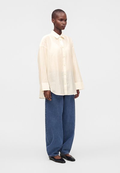 Personne aux cheveux courts portant une chemise crème boutonnée oversize, un jean bleu à jambes larges et des chaussures plates noires, debout devant un fond uni.