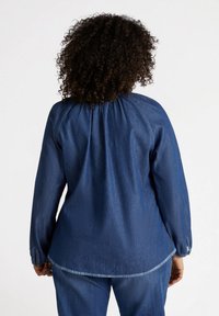 Blusa in denim blu scuro con scollatura arricciata e maniche lunghe. Presenta polsini elasticizzati e orlo curvo, mostrando un tessuto morbido e testurizzato.